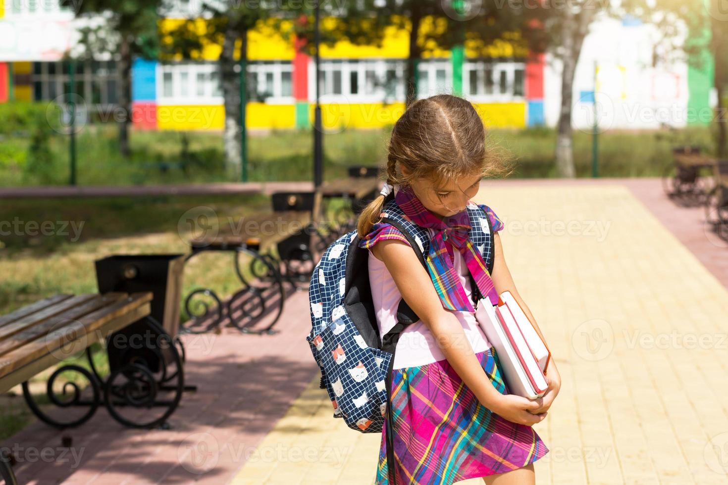 chica con mochila y una pila de libros cerca de la escuela. regreso a la escuela, el niño está cansado, pesados libros de texto. educación, clases de escuela primaria, el comienzo del año escolar, 1 de septiembre foto