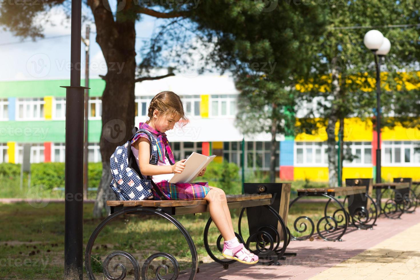 Girl with a backpack sitting on a bench and reading a book near the school. Back to school, lesson schedule, a diary with grades. Education, primary school classes, September 1 photo