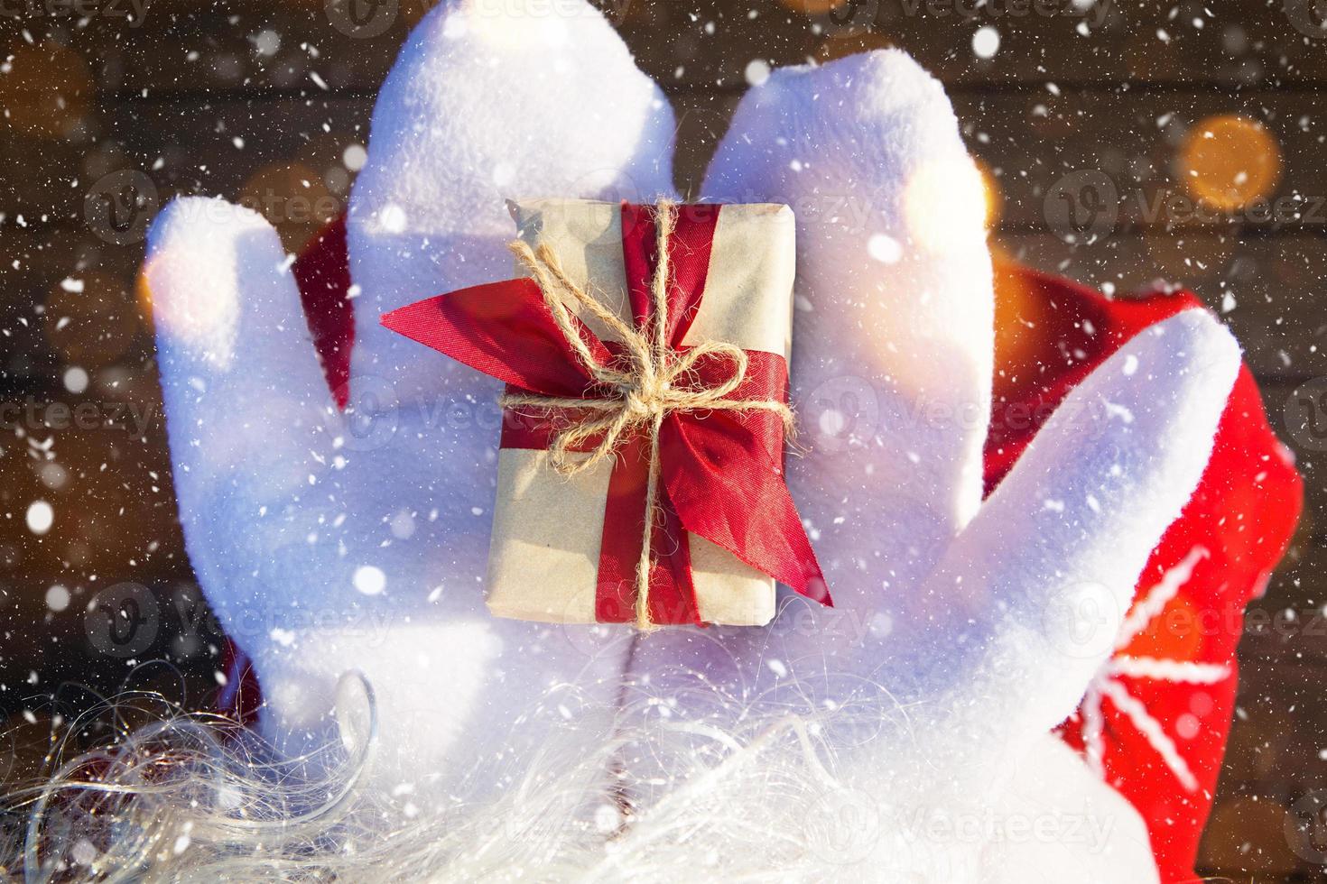 una caja con un regalo de navidad en manos de santa claus en mitones blancos. traje rojo, barba, guirnaldas de luces borrosas. año nuevo, preparación, expectativa de un milagro, un sueño hecho realidad. de cerca foto