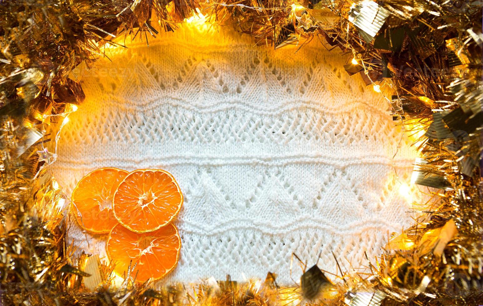 Festive frame made of gold garland with lights and tinsel, slices of dried oranges on a white knitted with a pattern background, copyspace. New year, Christmas, autumn and winter warming atmosphere. photo