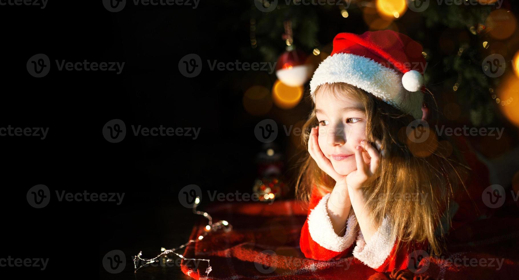 Little girl in a Santa hat and red dress under Christmas tree is dreaming, waiting for the holiday, lying on a plaid blanket. A letter on piece of paper, gifts. New year, Christmas. defocus lights photo