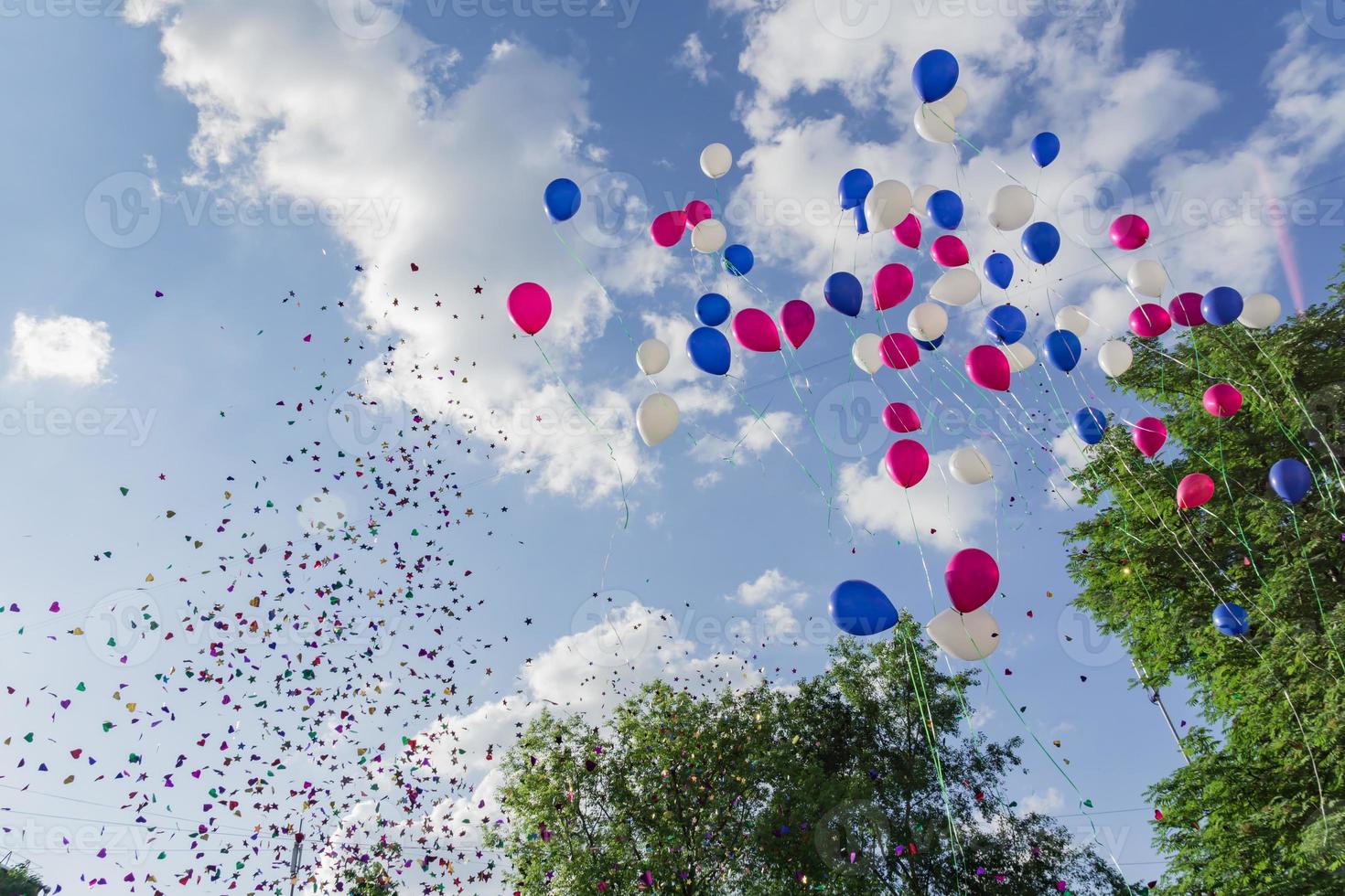 Multicolored balloons and confetti released in the blue sky against background of trees. Festive action photo