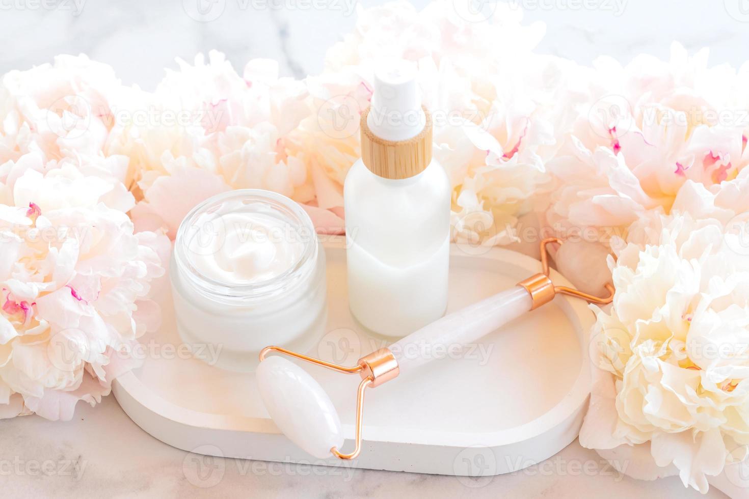 Facial kit for home skincare and spa. Rose quartz Face roller and jar of face cream on white marble background with peony flowers. Natural treatment concept. photo