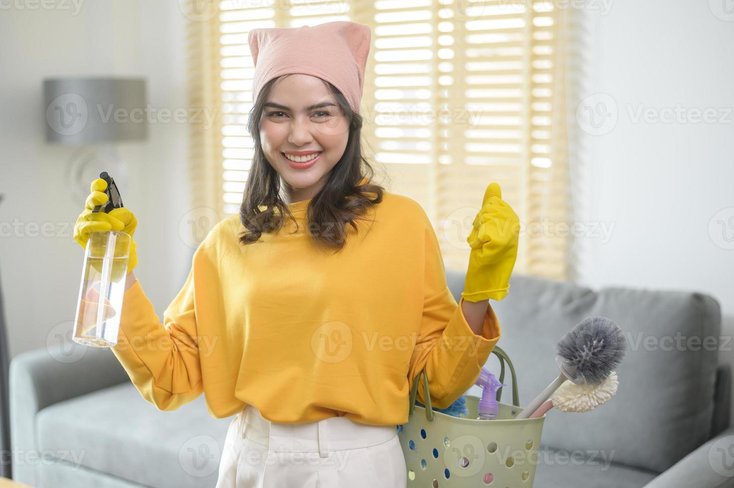 joven mujer feliz usando guantes amarillos y sosteniendo una canasta de artículos de limpieza en la sala de estar. foto