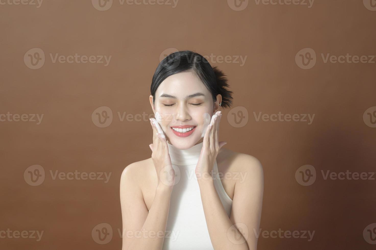 A young woman with beautiful face smiling, washing her face over brown background , beauty skin care concept photo