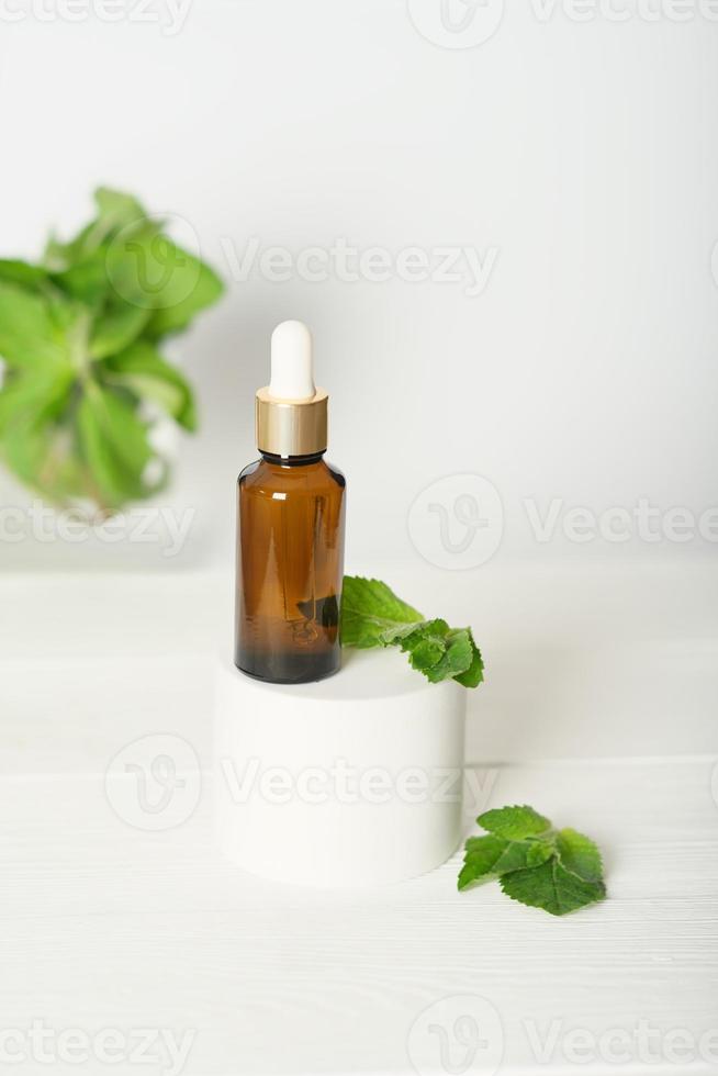 maqueta de una botella cuentagotas de vidrio sobre un podio blanco sobre un fondo blanco, con hojas de menta. producto de suero cosmético sobre un fondo blanco. vertical con hojas de menta foto