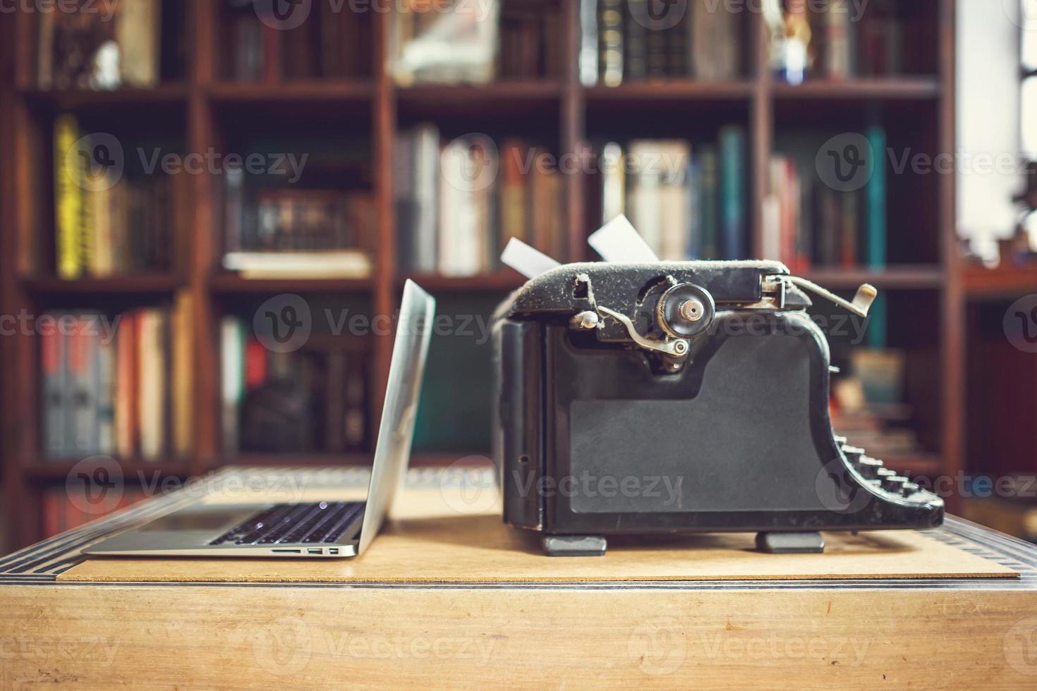 máquina de escribir antigua cubierta de polvo con una hoja de papel blanco cerca de un portátil moderno en el fondo de la librería. tecnología moderna y electrodomésticos antiguos foto