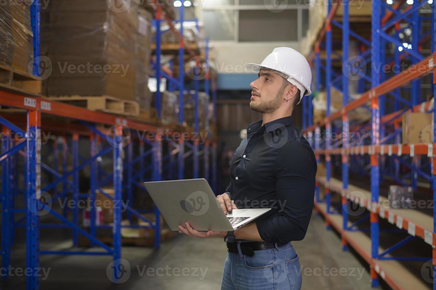 joven trabajador caucásico con casco usando una computadora portátil en un almacén moderno. foto