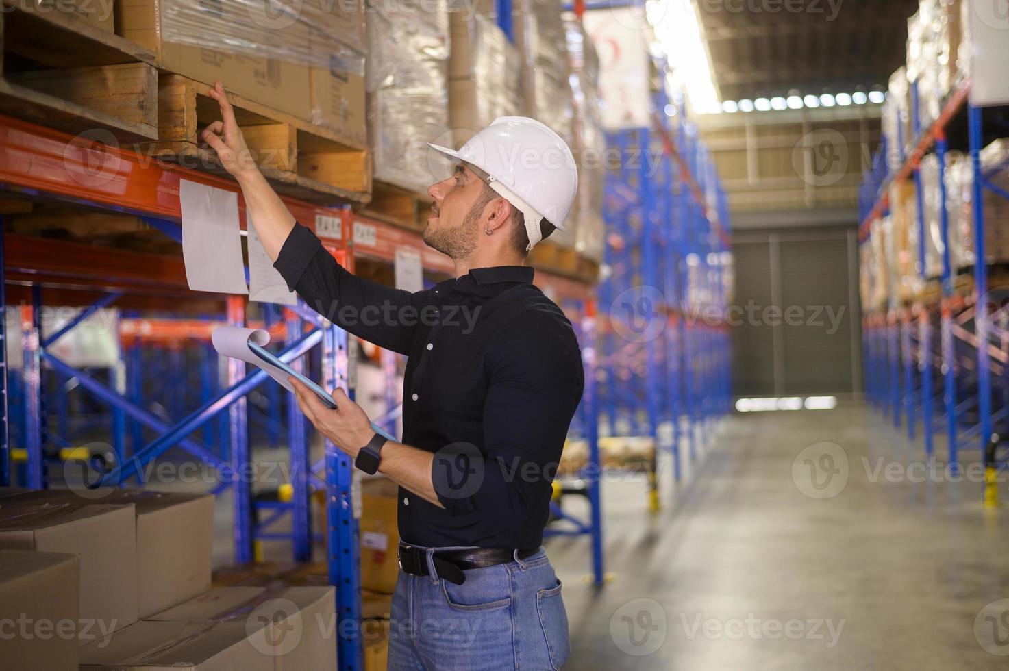 Young worker wearing helmet checking inventory and counting product on shelf in modern warehouse. photo