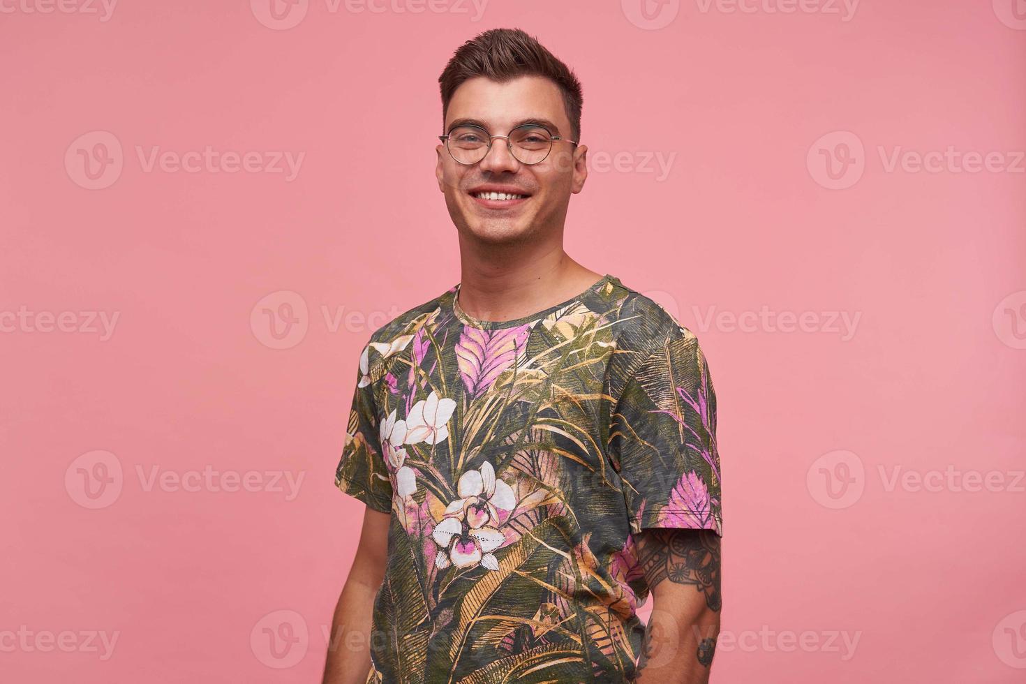 Studio shot of beautiful young tattooed male with charming smile posing over pink background, looking happily to camera, wearing glasses and flowered t-shirt photo