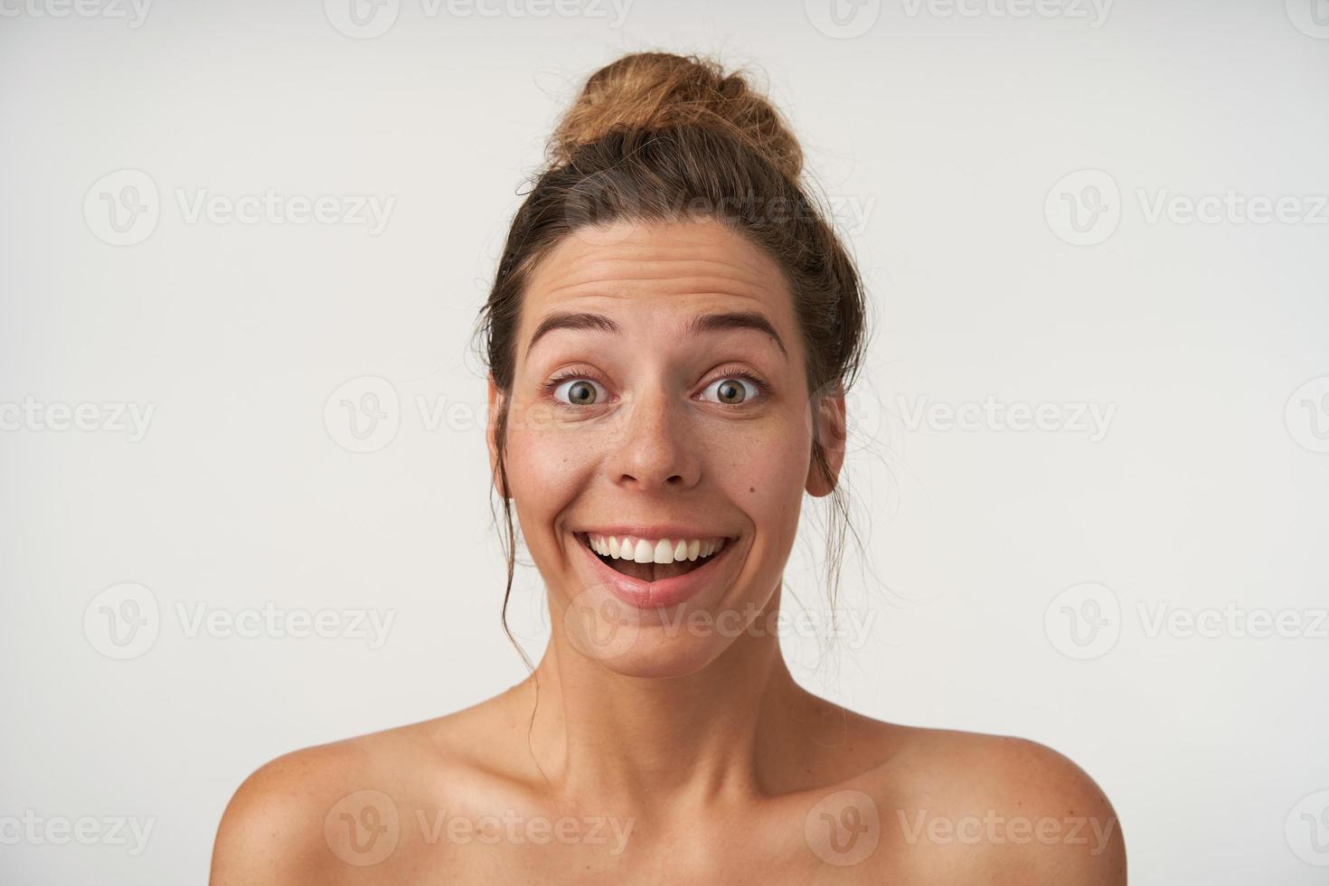 Indoor close-up of surprised young pretty woman with casual hairstyle standing over white background, looking to camera with raised eyebrows and amazed face photo