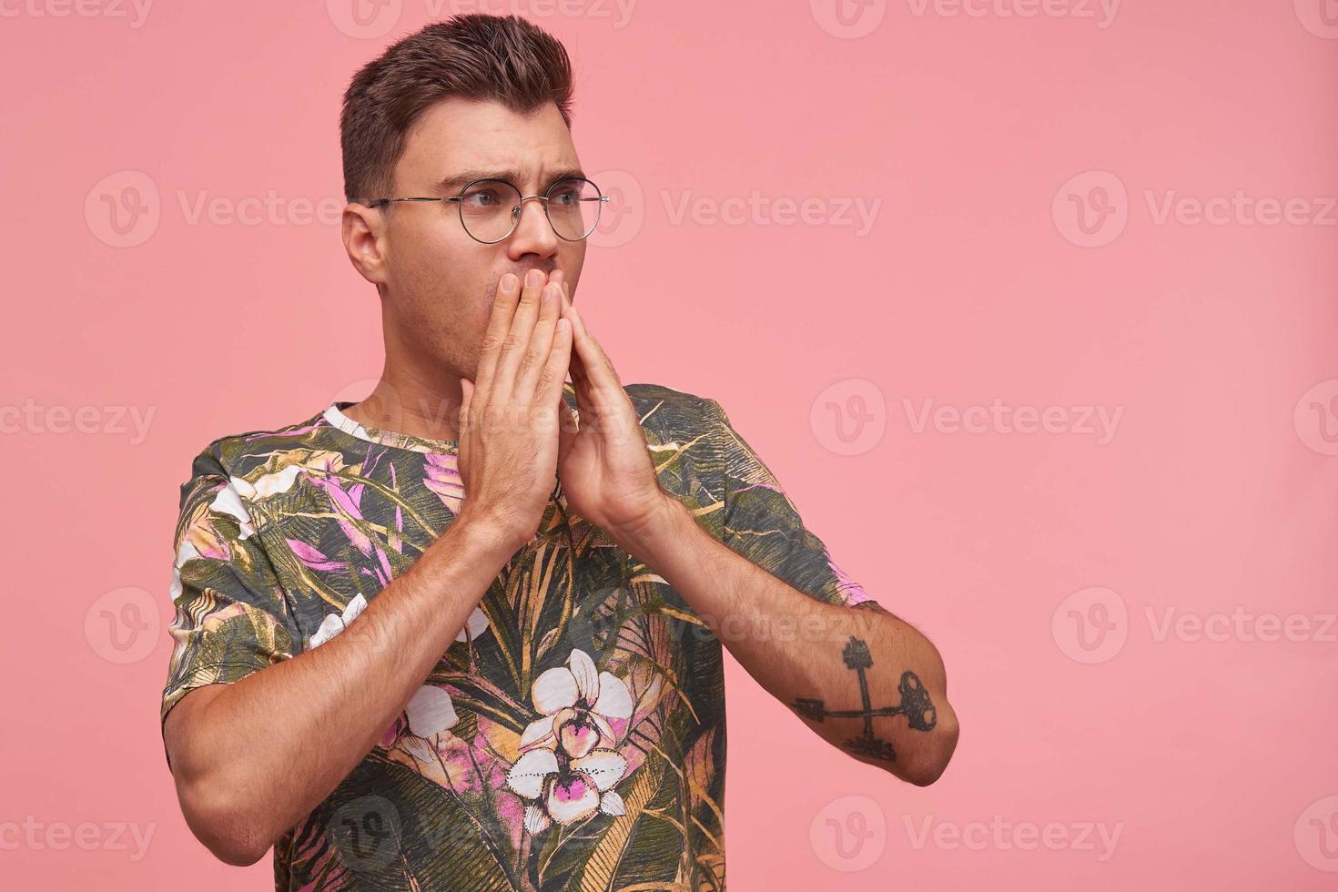 Portrait of amazed young man with glasses in casual t-shirt astonishing and covering mouth with both hands, looking with stunned expression aside, isolated over white background photo