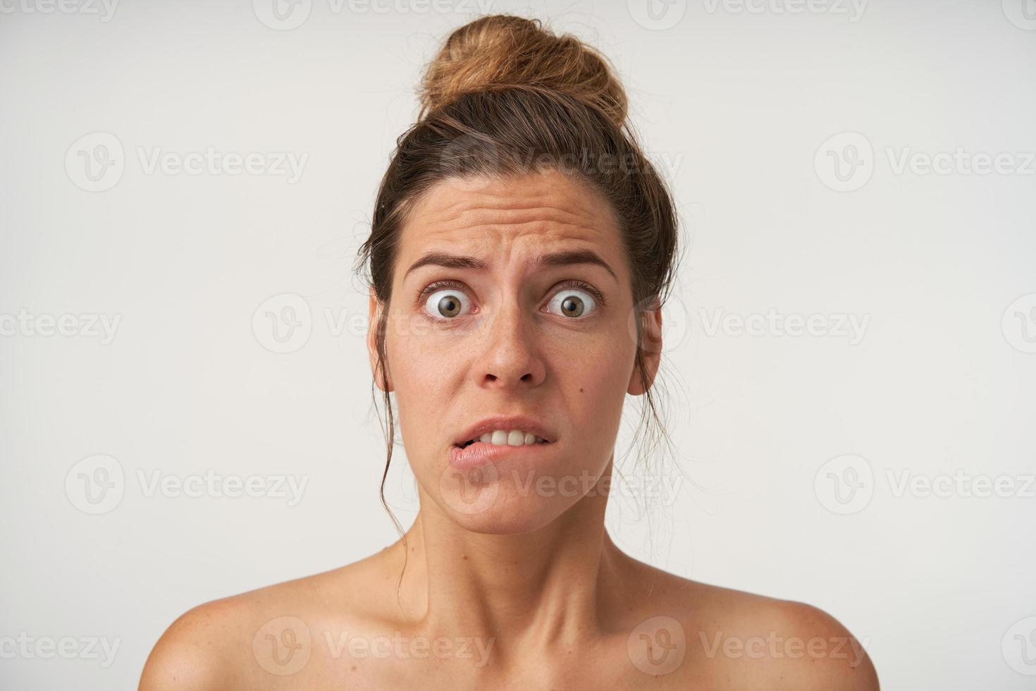 Indoor shot of young frightened woman with bun hairstyle, wrinkling forehead and biting underlip, looking to camera with wide eyes opened photo