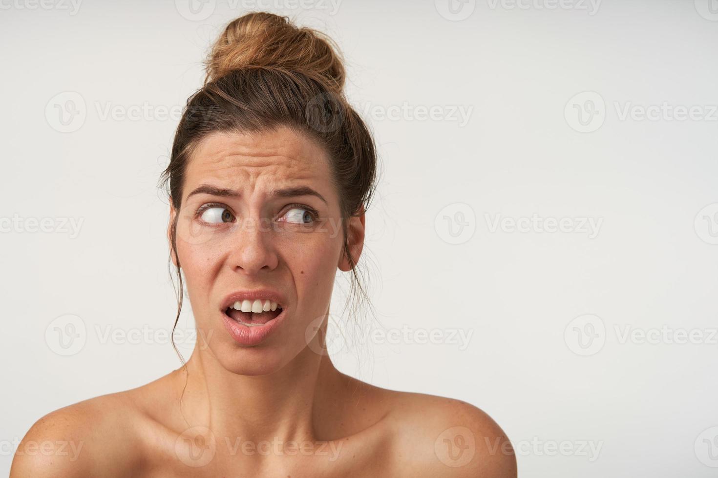 Head shot portrait of shocked frustrated young female with bun hairstyle, looking aside with astonished grimace, isolated over white background photo