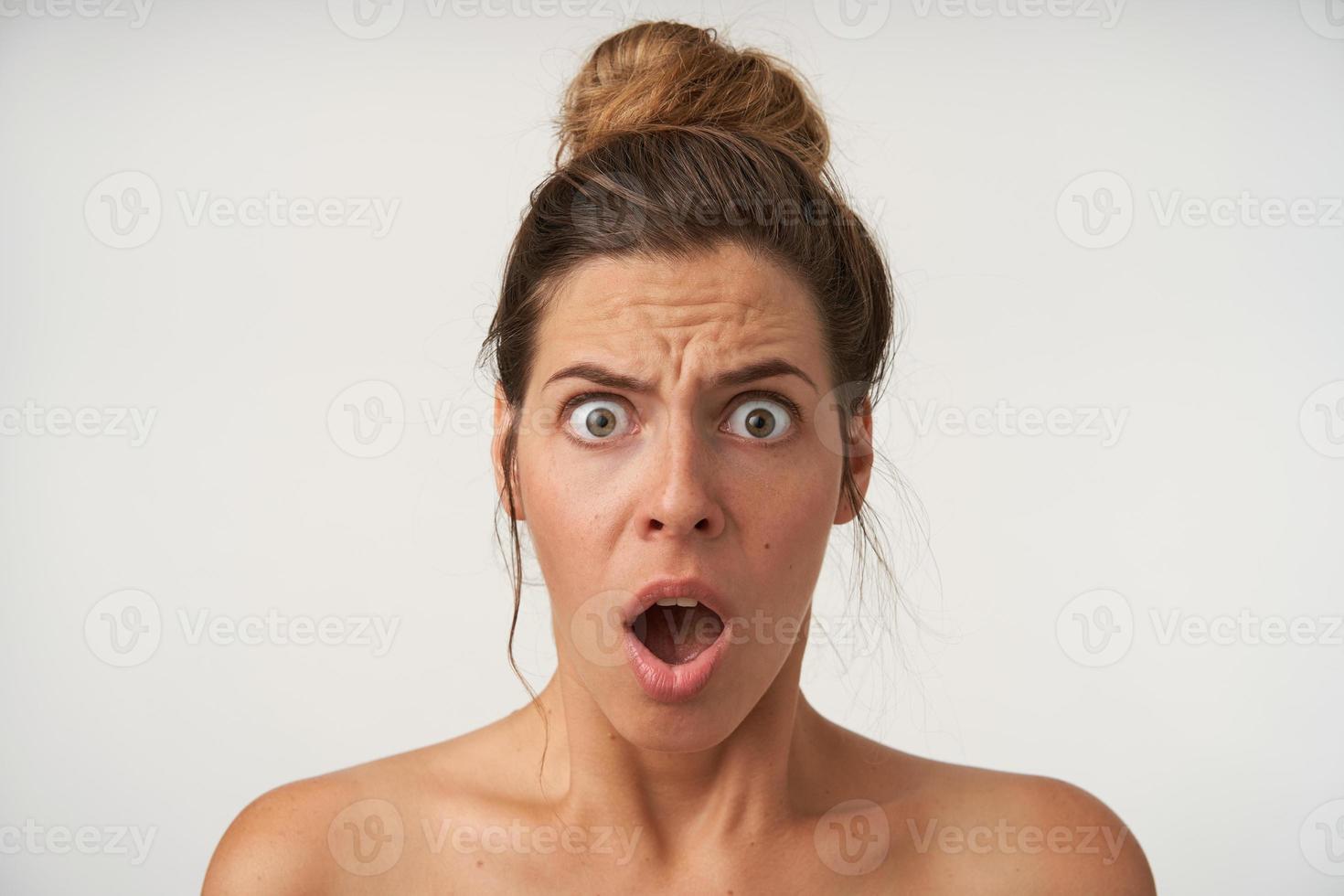 Indoor portrait of shocked young female with casual hairstyle, posing over white background with surprised face, looking to camera with wide opened mouth and eyes photo