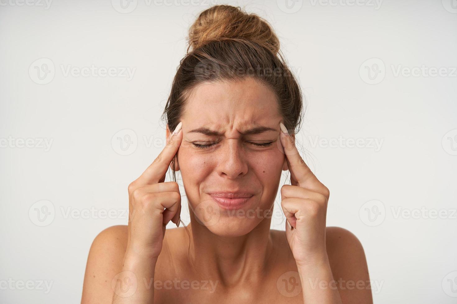 Close-up of young pretty woman with painful face, posing over white background with index fingers at temples, closing eyes because of headache photo