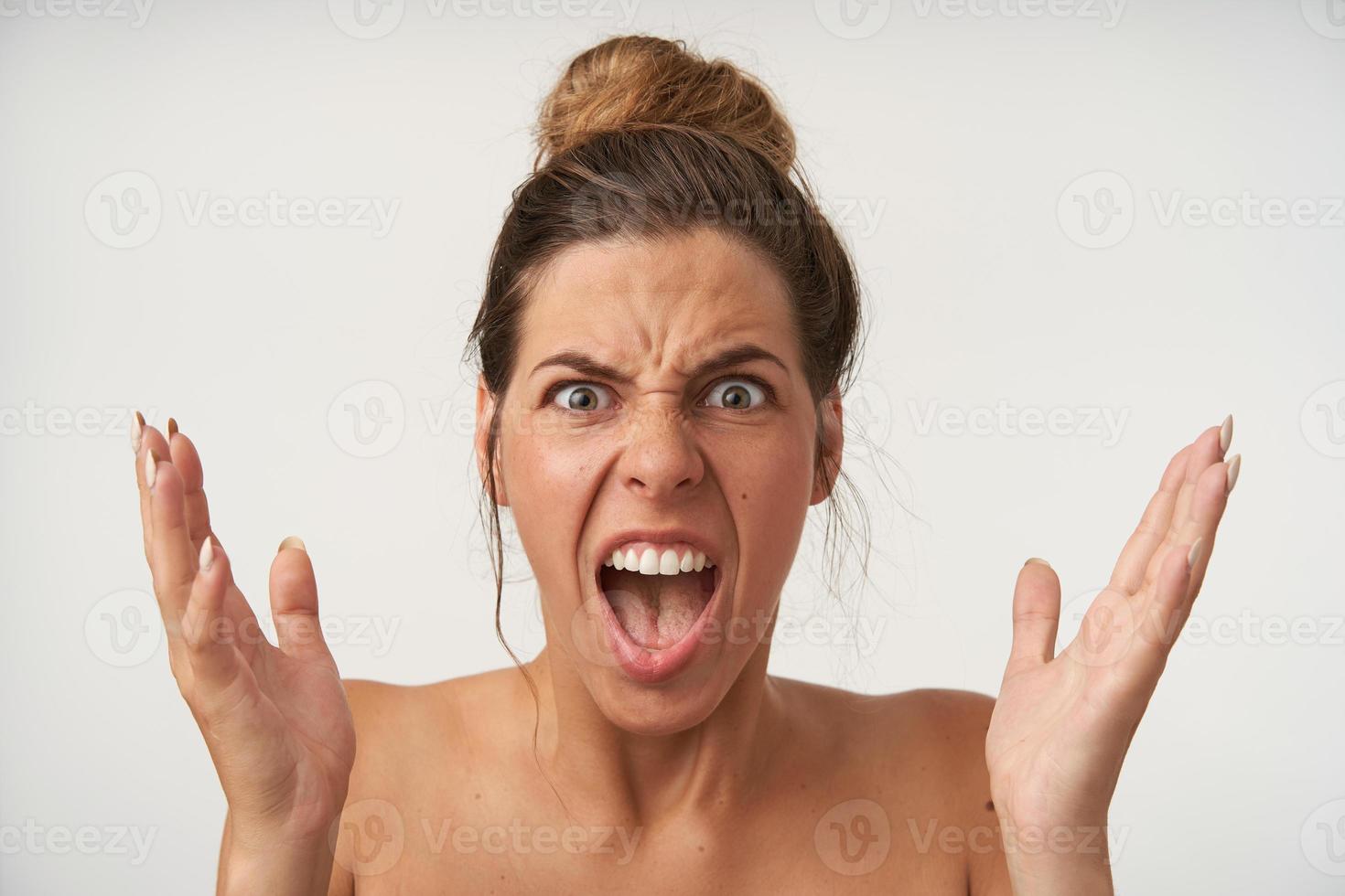Close-up of wrathful young lovely woman posing over white background with raised palms, frowning and shouting with wide mouth opened photo