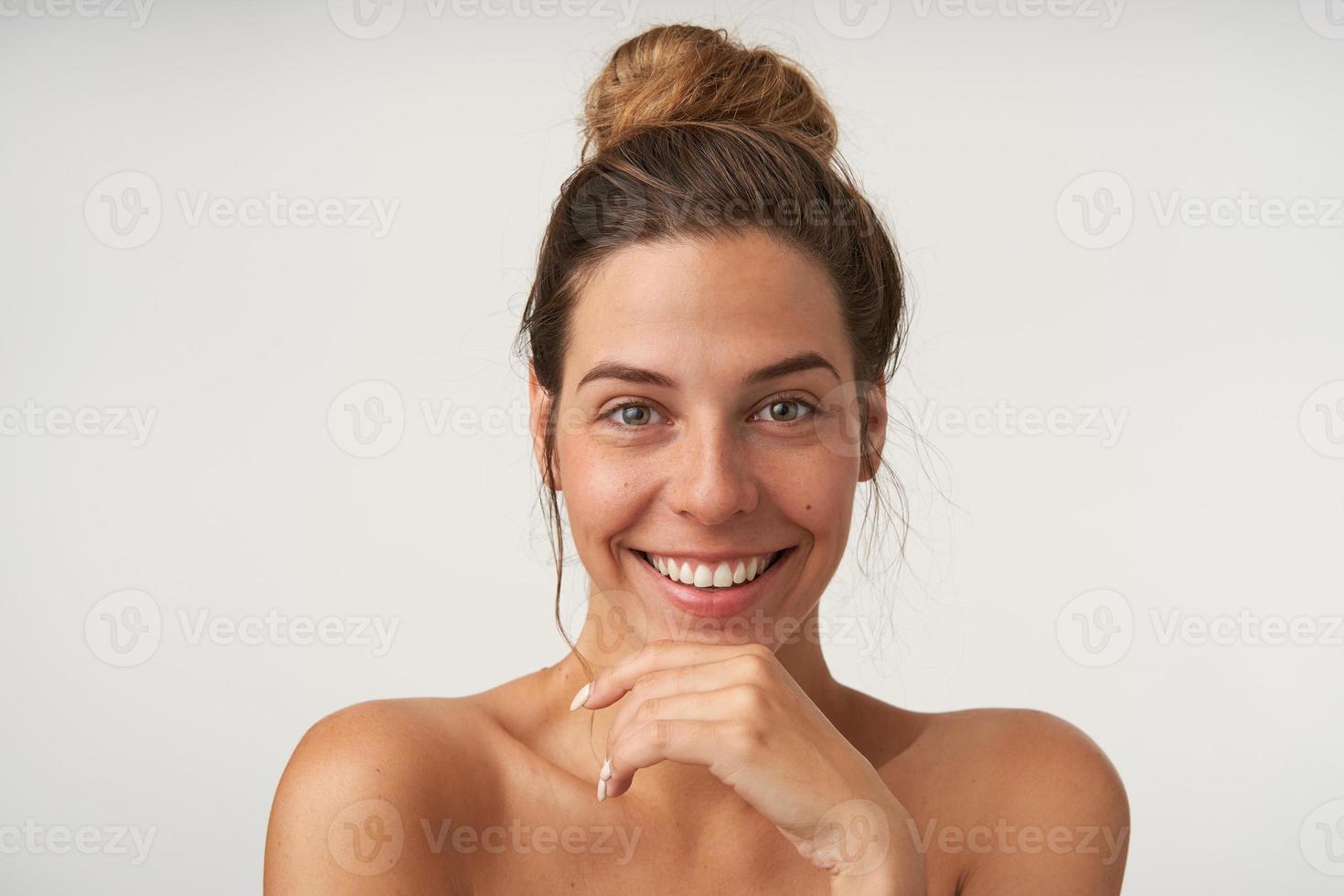 Good looking young happy female posing over white background with charming smile, wearing bun hairstyle and no make-up, keeping hand on her chin photo