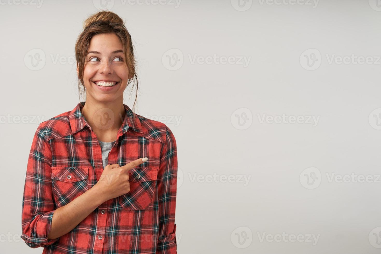 Happy young lovely woman with bun hairstyle posing over white background without make-up, pointing with index finger aside, smiling widely and being in nice mood photo