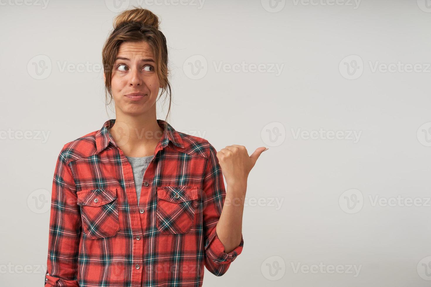 Indoor portrait of young lovely female with bun hairstyle posing over white background, looking aside with doubting, tumbing aside with hand photo