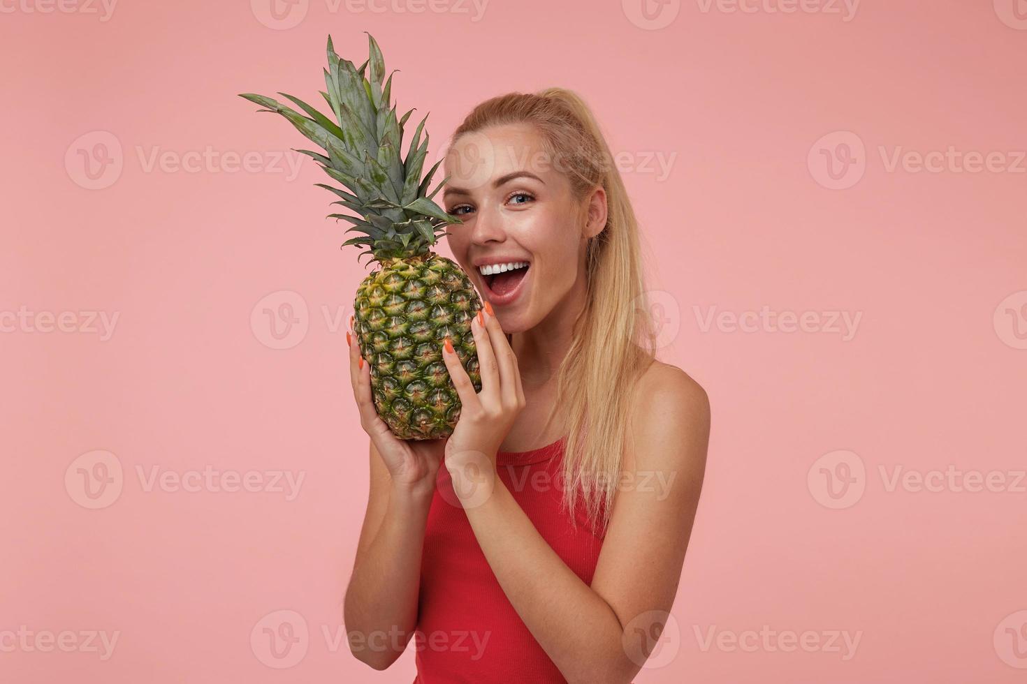 Studio shot of young blonde female with casual hairstyle holding pineapple in her hands, going to bite pineapple, looking at camera and smiling joyfully photo