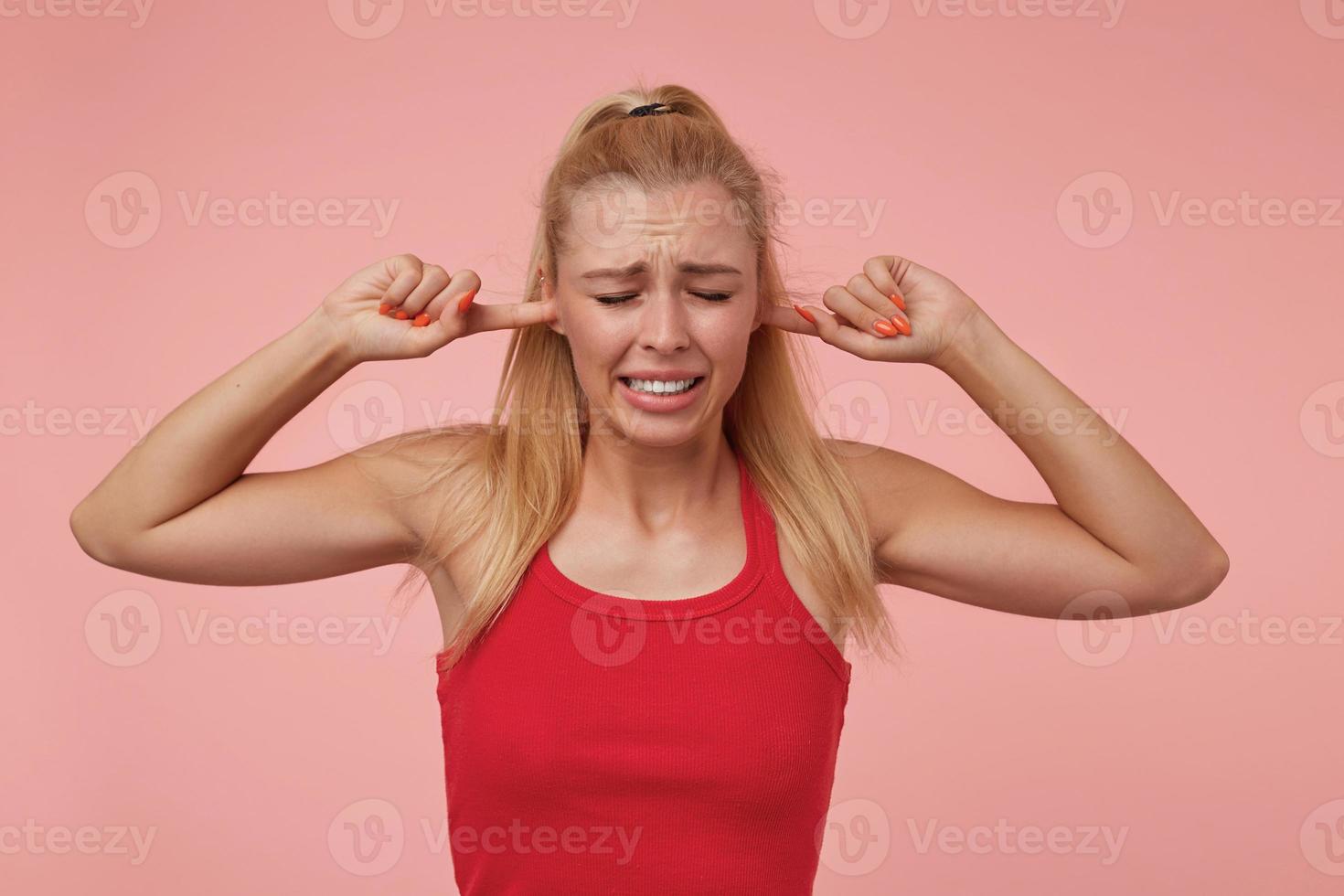 Studio photo of good looking young woman with casual hairstyle standing over pink background, covering ears with hands and wrinkling, trying to avoid loud sounds