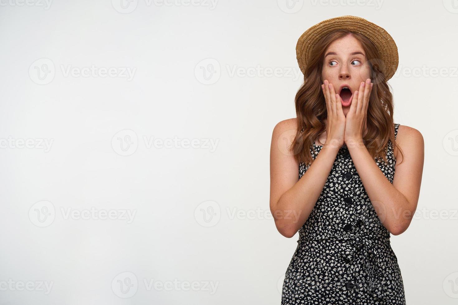 Studio photo of amazed beautiful red haired woman in boater hat looking aside with wide eyes and mouth opened, holding palms on her face and raising eyebrows, isolated over white background