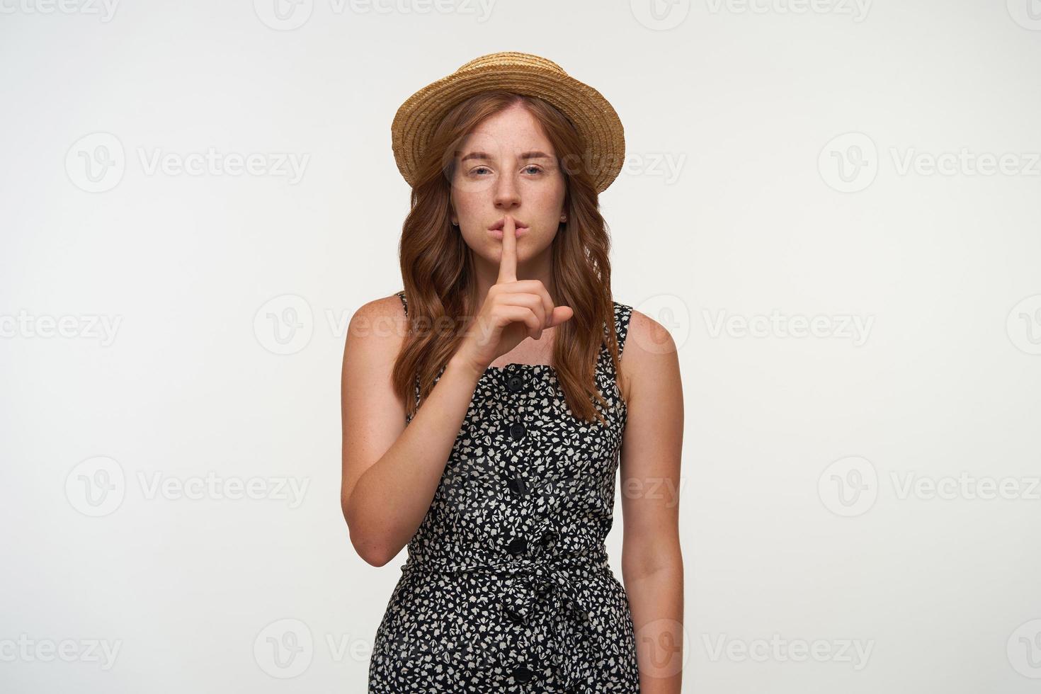 Attractive young red haired lady in romantic dress and straw hat posing over white background, raising index finger to lips, asking to keep secret photo