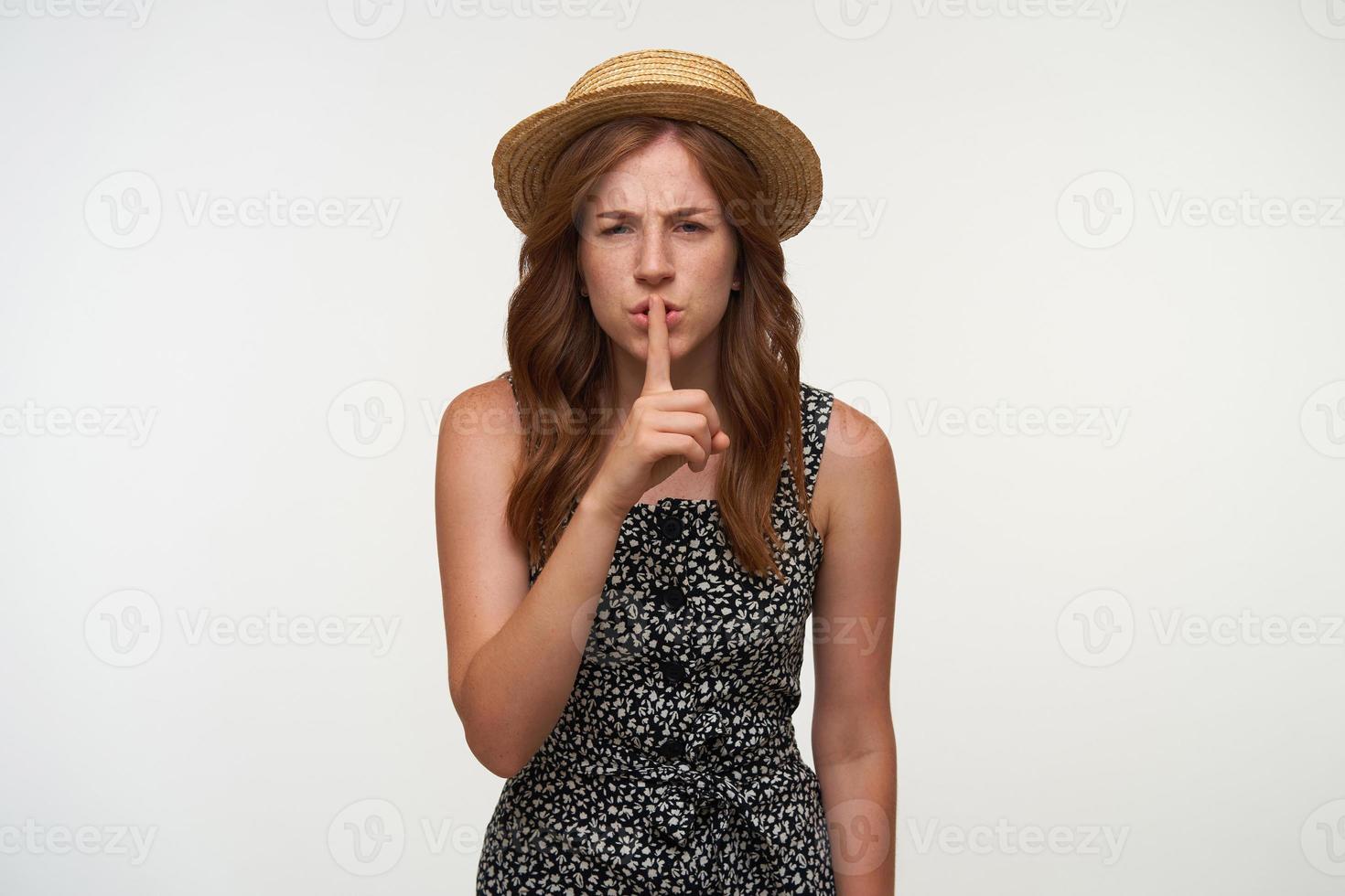 Irritated young red haired woman wearing casual dress and boater hat, making hush gesture, asking to be quiet, frowning and looking to camera seriously photo