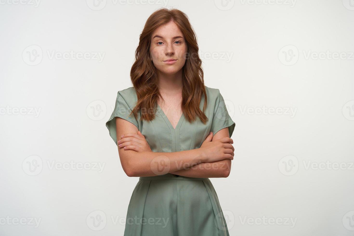 Portrait of pretty redhead female in pastel dress looking to camera with mysteriously smile, standing over white background with crossed arms photo