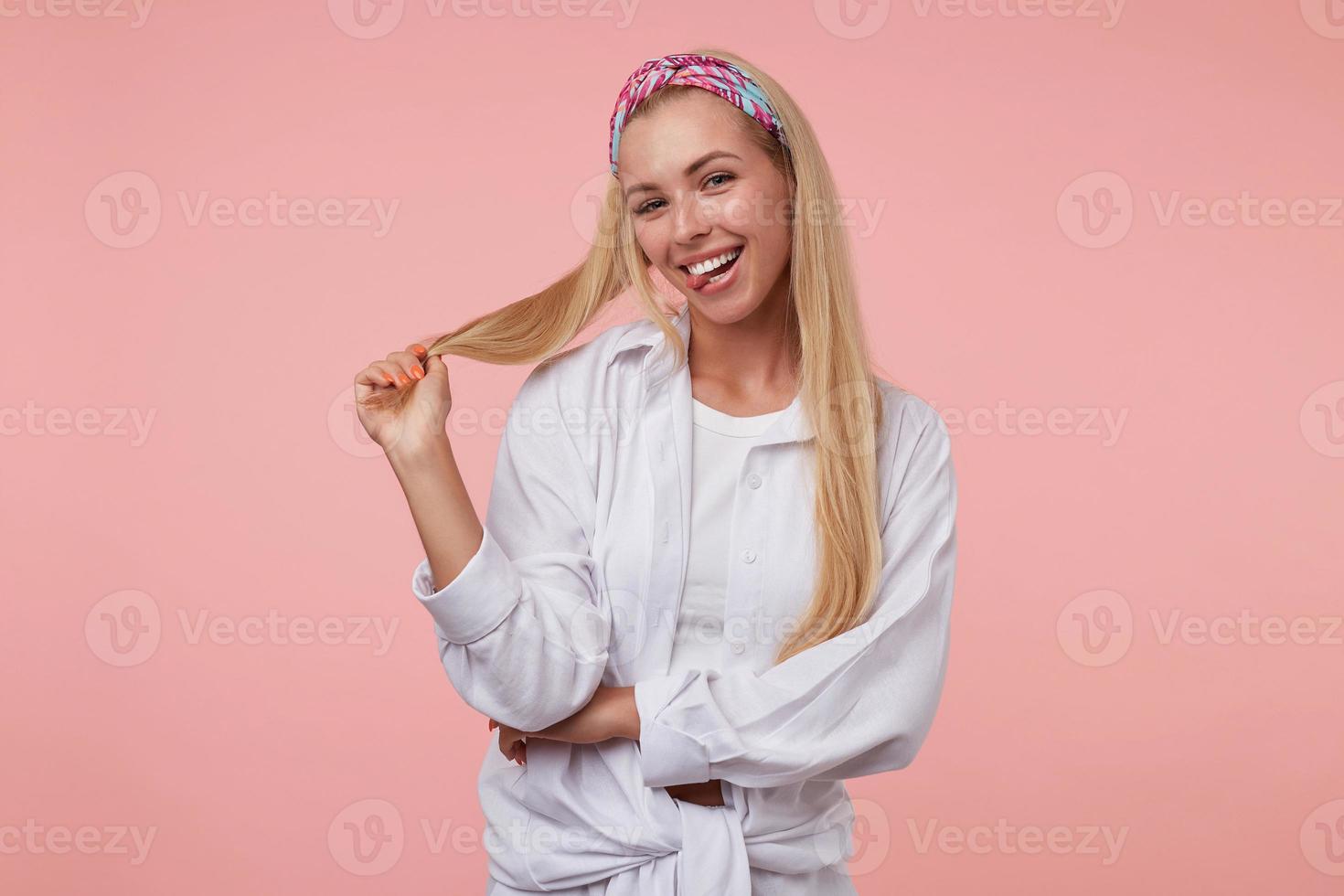 primer plano de una encantadora mujer rubia mirando juguetonamente a la cámara y tirando de su cabello, sonriendo y mostrando la lengua, de pie sobre el fondo rosa foto