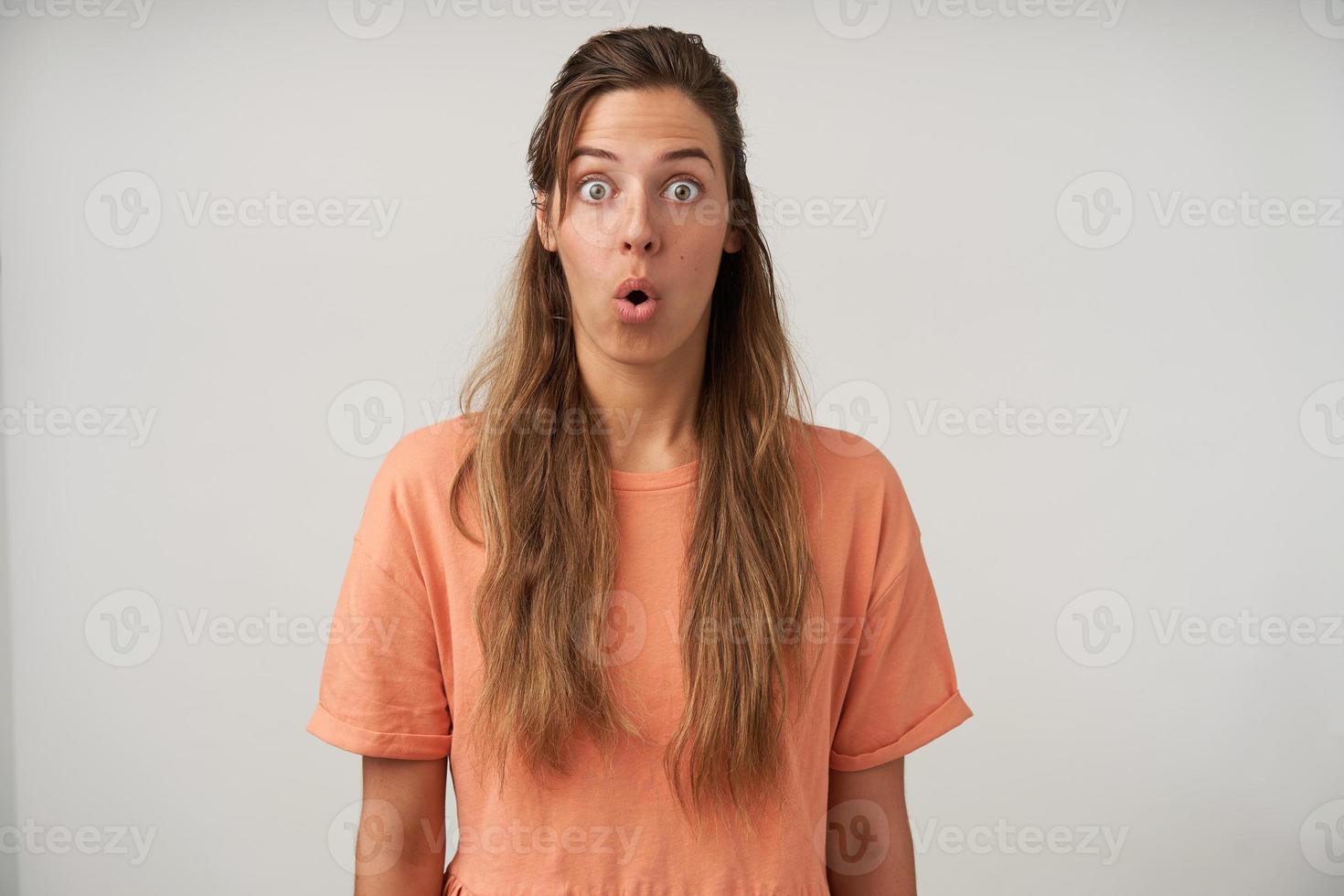 Indoor photo of surprised young woman with long hair, looking to camera with wide eyes opened and folded lips, wearing peach t-shirt, isolated over white background