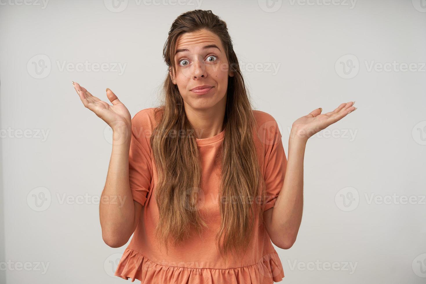 retrato de una joven mujer de pelo largo encogiéndose de hombros con las manos extendidas de lado y sonriendo, usando una camiseta de melocotón, sin tener idea de cómo resolver el problema foto