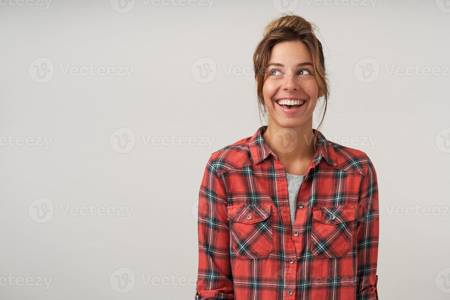 Happy young woman with casual hairstyle posing over white background in checkered shirt, looking aside cheerfully with broad sincere smile photo