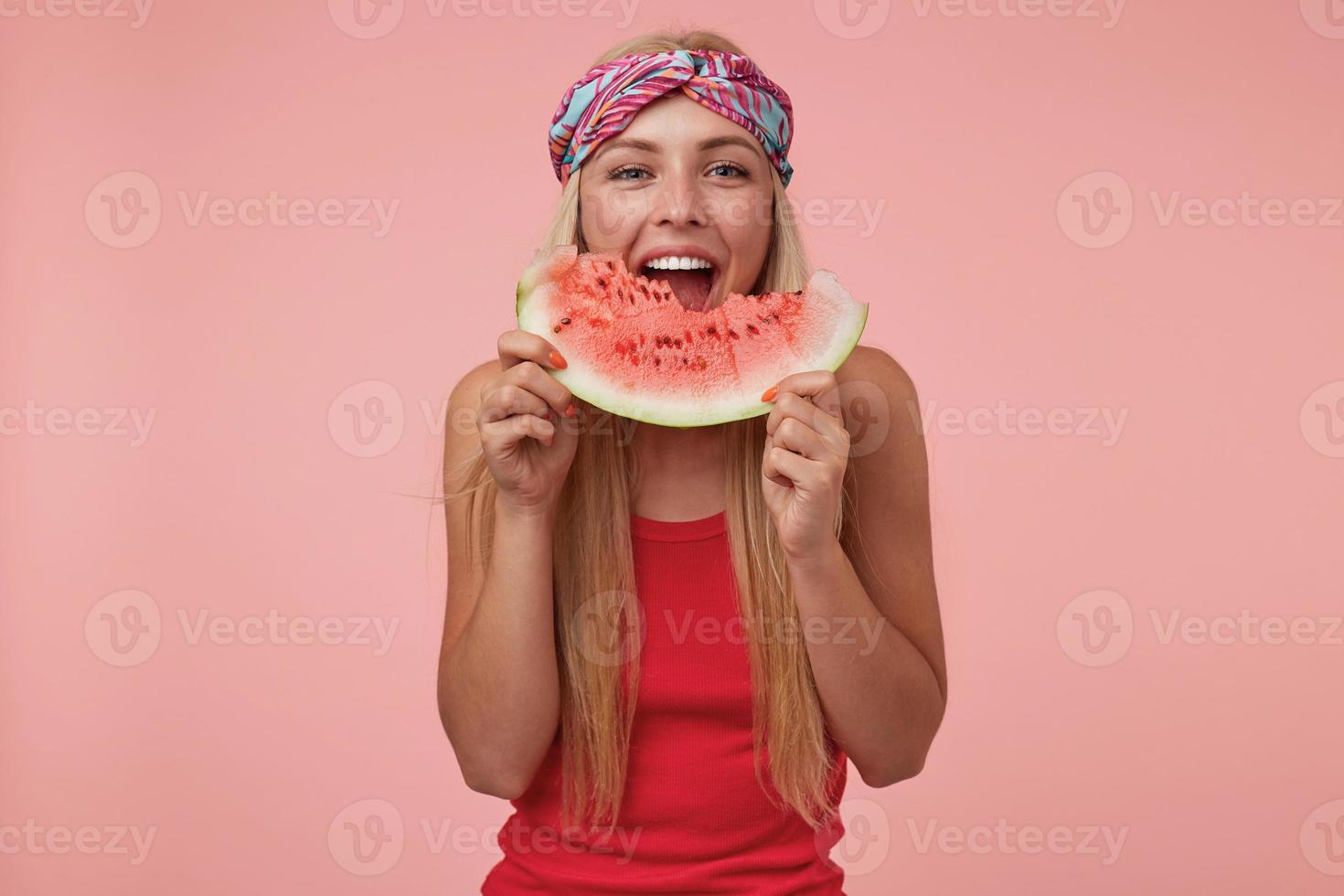 Indoor shot of joyful pretty female with headband and long blonde hair posing over pink background, eating watermelon, being in nice mood photo