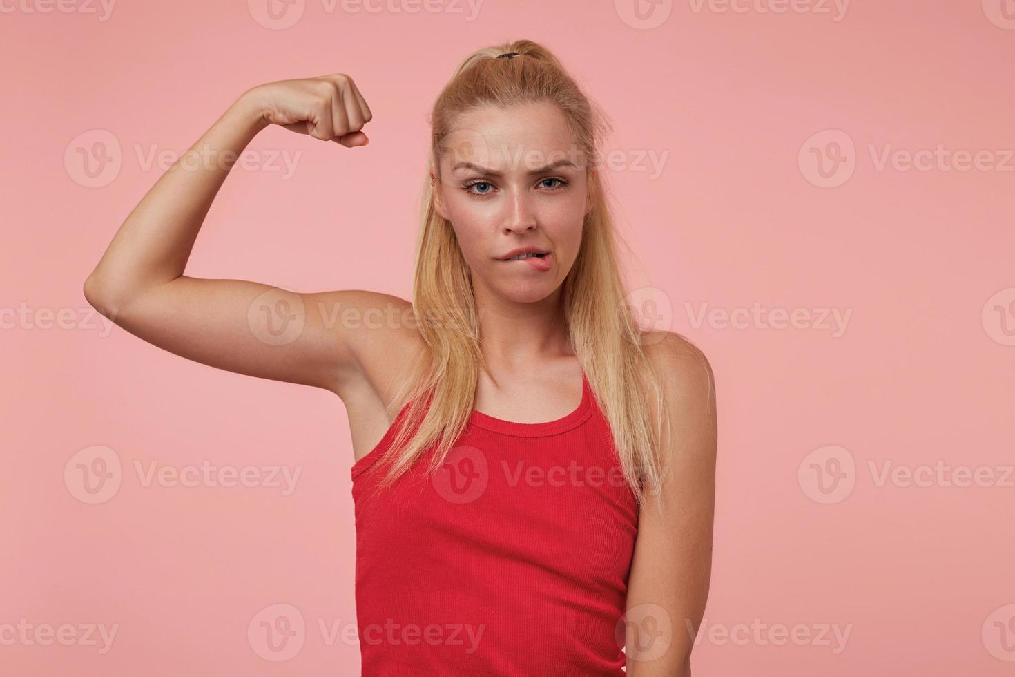 Indoor photo of attractive young lady looking at camera menacingly, showing her pumped up hand, playing with eyebrows and biting her lip
