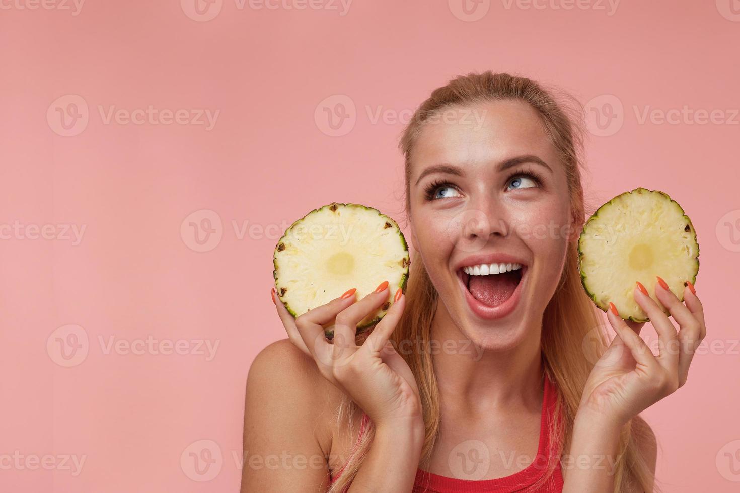 Happy young attractive female with natural make-up and casual hairstyle looking joyfully upwards, having fun with rings of fresh pineapple, isolated over pink background photo