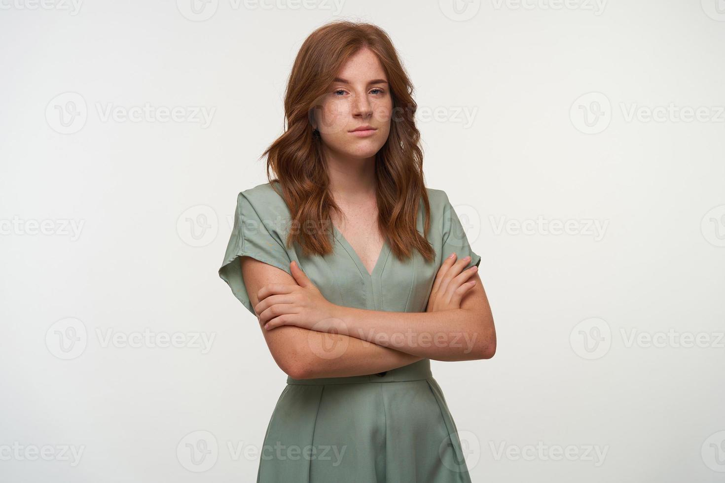 Indoor shot of pleasant looking young female with romantic hairstyle, crossing hands on her chest with calm face, wearing pastel dress, isolated over white background photo