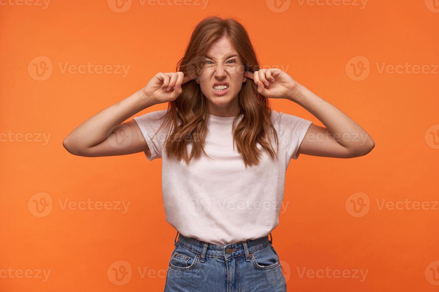 Studio photo of beautiful curly female in casual clothes grimacing and puckering face, closing ears with forefingers, trying to avoid loud sounds