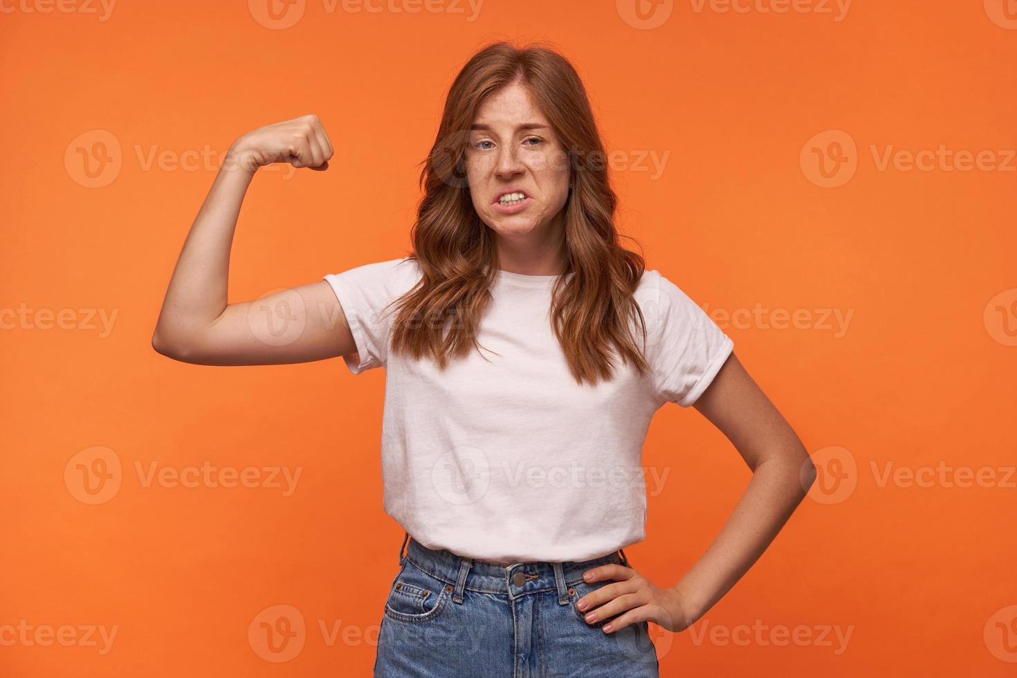 Studio shot of pretty redhead curly young woman showing her power in raised hand, wearing casual clothes, posing over orange background photo