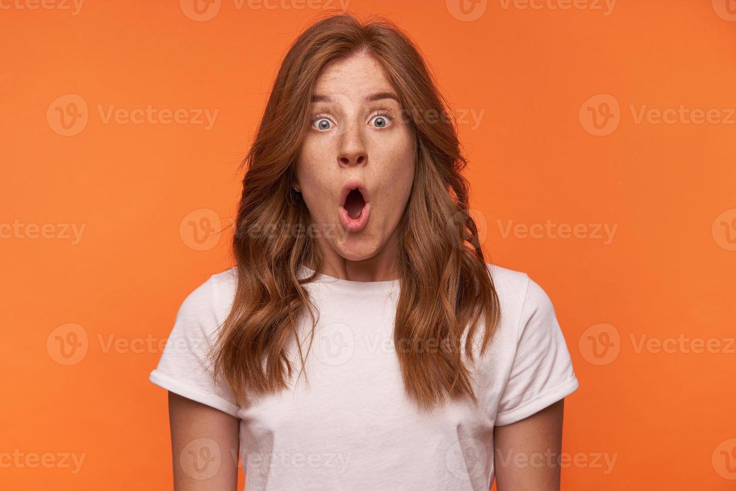 Close-up portrait of shocked curly female in white t-shirt posing over orange background with open-eyed face, raising eyebrows and opening mouth widely photo