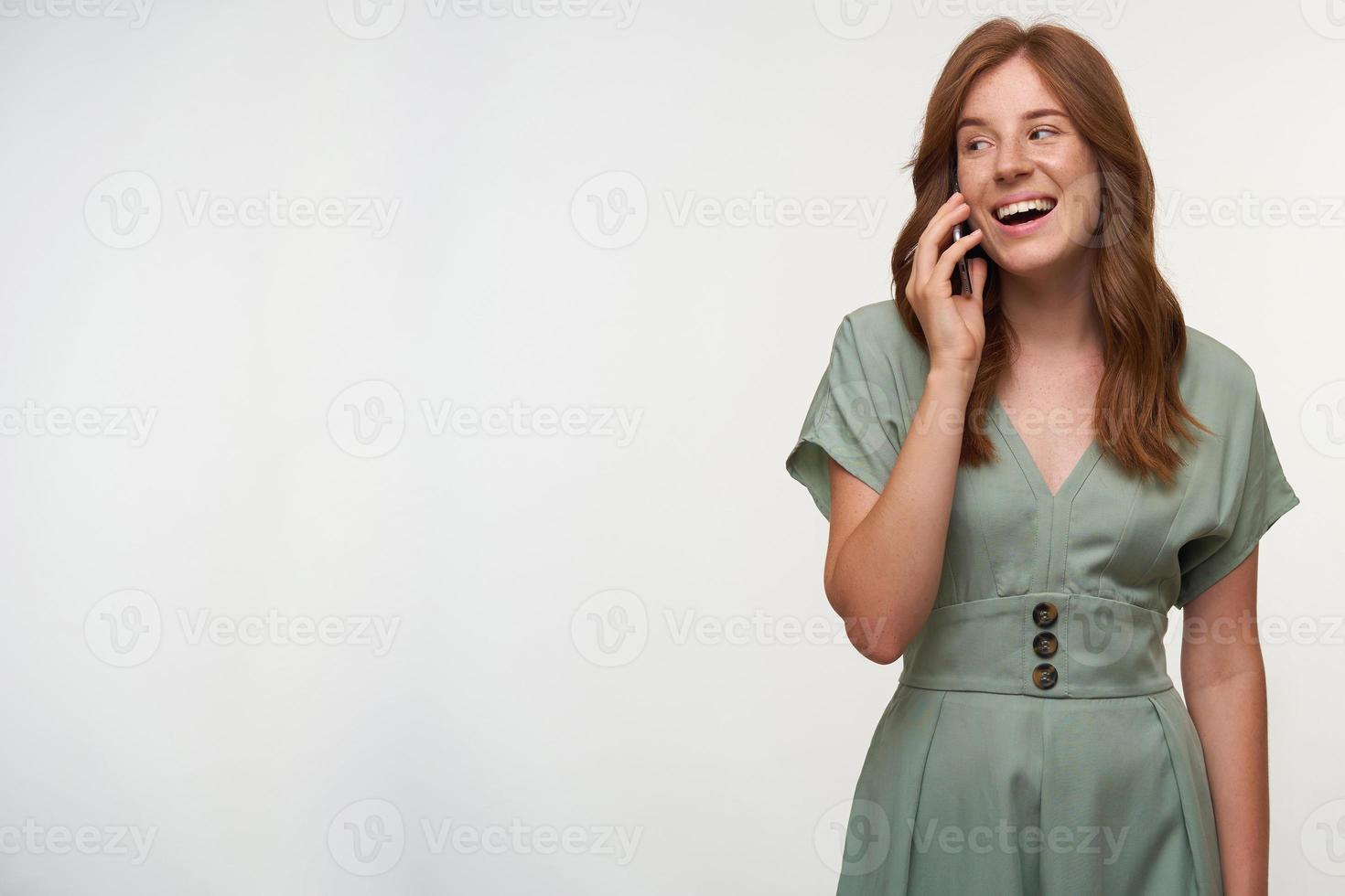 Indoor shot of happy young redhead woman in pastel dress posing over white background, looking aside and smiling widely, having pleasant talk on phone photo