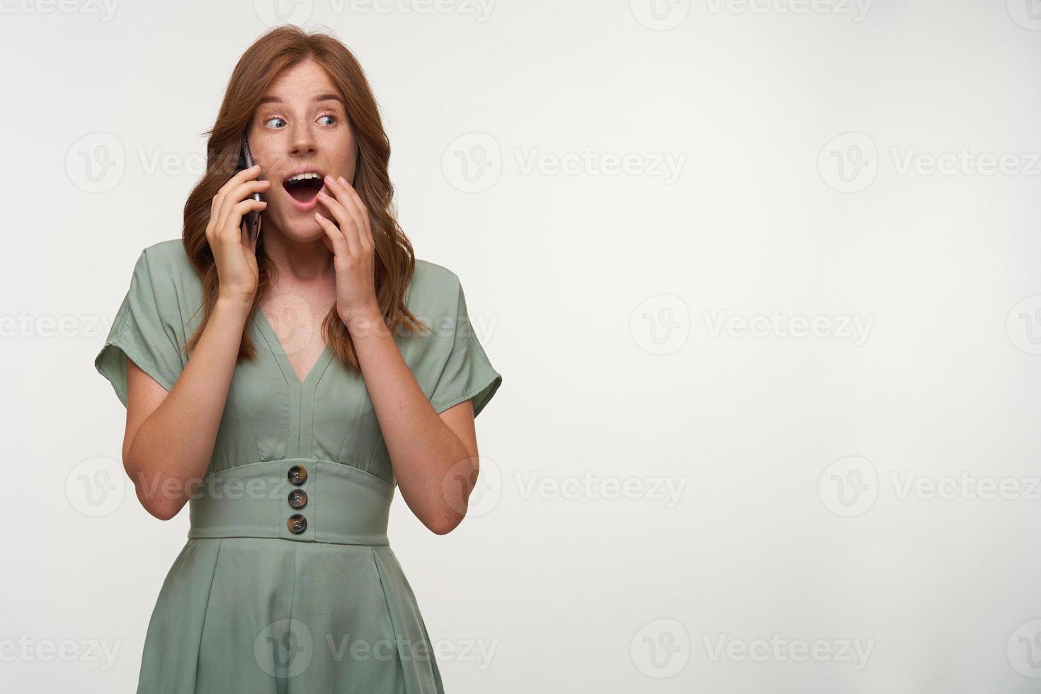 Amazed young beautiful female with red hair posing over white background, opening mouth widely and covering it with hand, holding smartphone by her ear photo