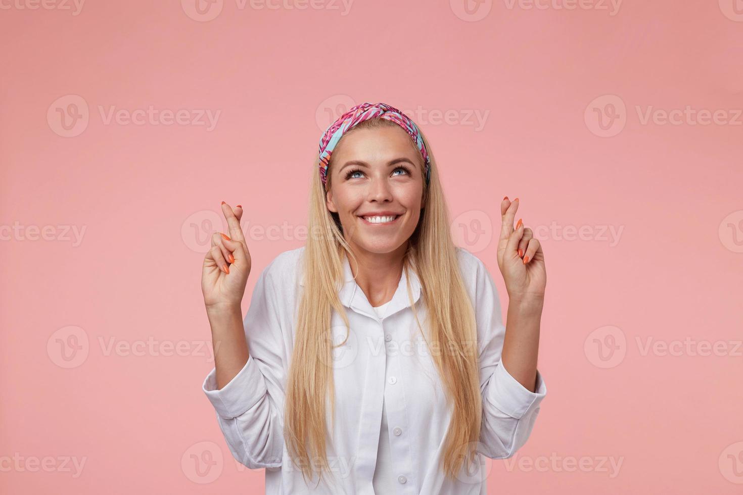 Indoor shot of good looking blonde woman crossing fingers and believing in better, looking hopefully upwards, has shining smile and eyes full of faith, isolated over pink background photo