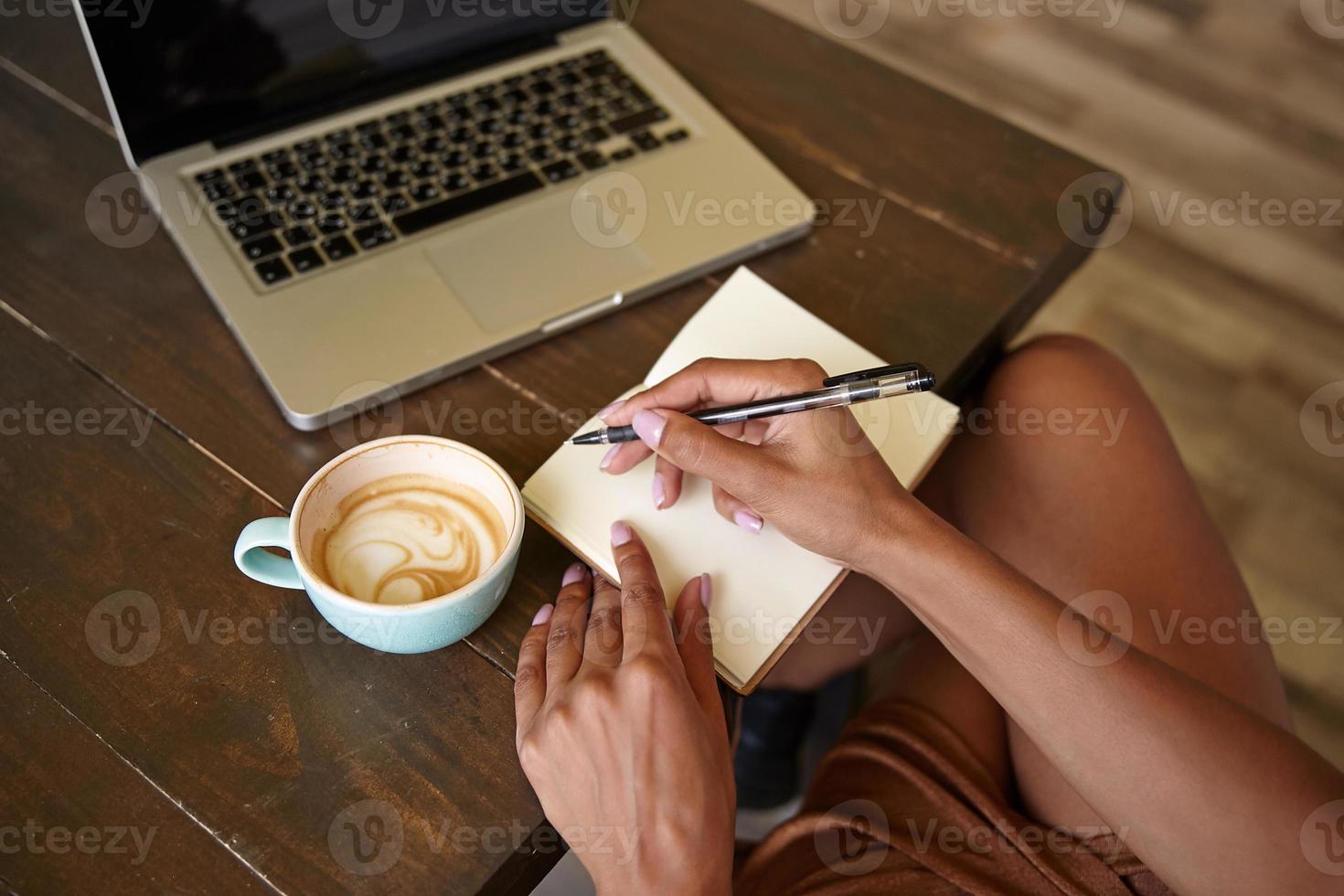 Indoor close-up of wooden countertop with laptop on it, freelancer female working in public place with her notebooks and drinking coffee photo