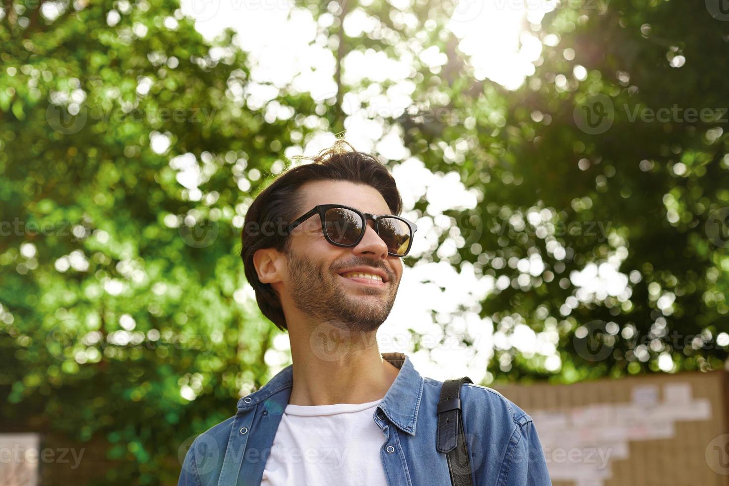 Outdoor portrait of happy pretty bearded male with dark hair looking aside and smiling cheerfully photo