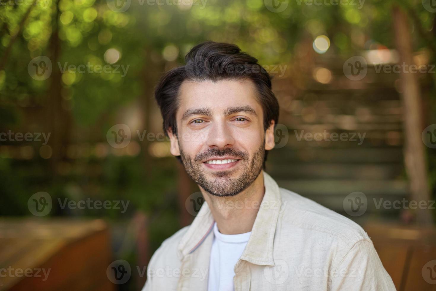 Close-up of beautiful bearded male with dark hair posing over green ciy park on sunny day photo