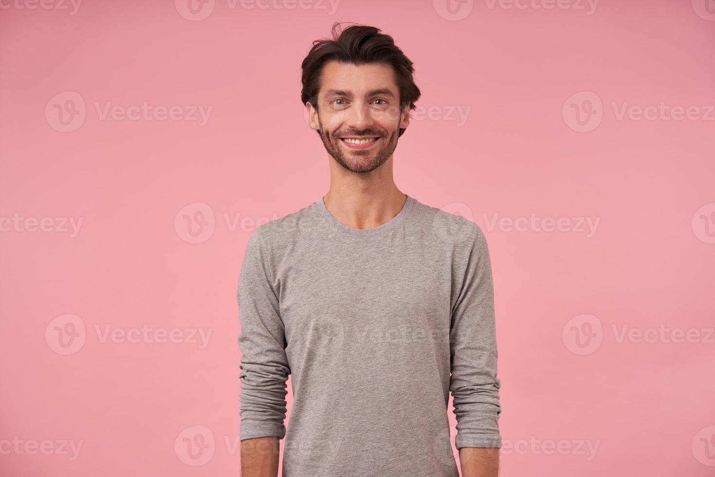 Studio shot of beautiful bearded male with dark hair standing over pink background, wearing casual clothes, looking at camera cheerfully with hands down photo