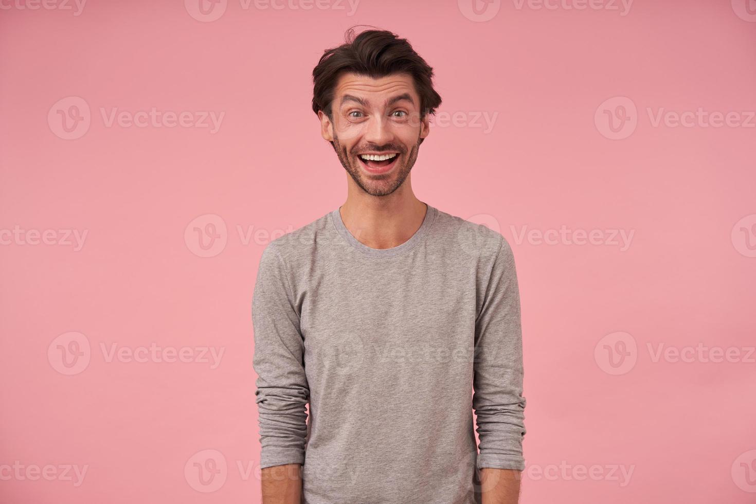 retrato interior de un hombre barbudo feliz con un corte de pelo moderno con ropa informal, posando sobre un fondo rosa con las manos hacia abajo, mirando la cámara alegremente con la boca abierta foto