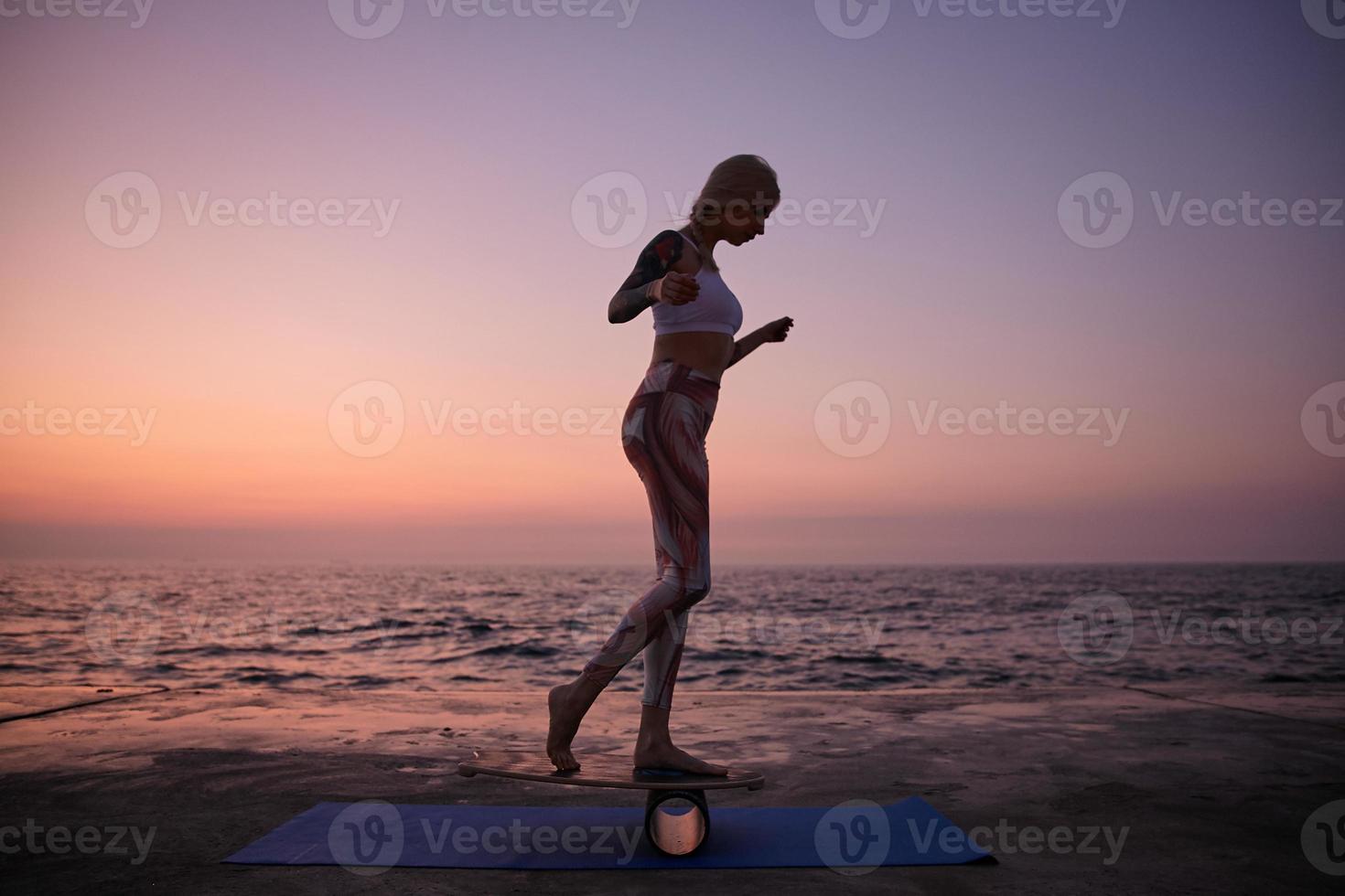 Outdoor photo of sporty slim female with casual hairstyle in sporty wear standing on balance board, doing sports with balancer on seafront during sunrise, raising hands to save equilibration